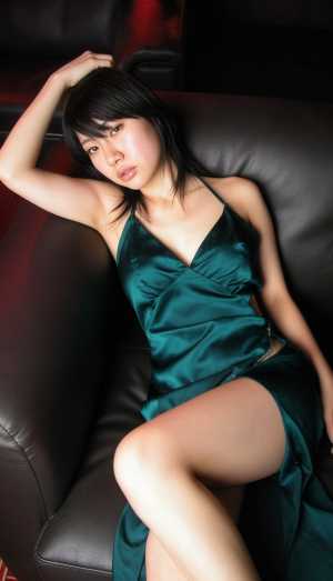 ティールのサテンのドレスを着て、黒い革張りのソファでくつろぎ、自信に満ちたポーズをと...。