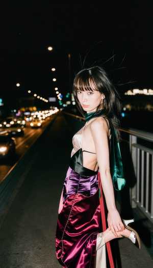 紫と黒のサテンの着物を着て首に緑のスカーフを巻いて、夜の橋の上に立つ若い女性をサイド...。