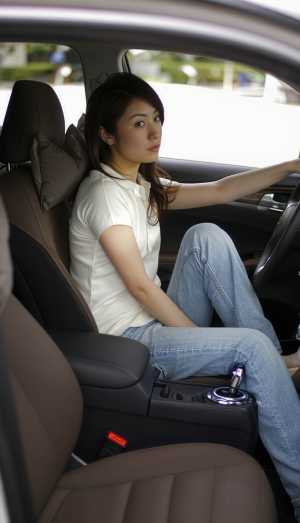 白いブラウスとブルー ジーンズを着て、現代の車の運転席に座っている若い女性を横から撮...。