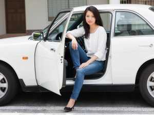 白いブラウスとブルー ジーンズを着て、白い車の運転席に座っている若い女性を横から撮影...。