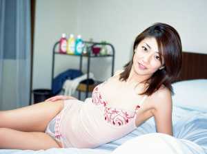 赤い刺繍が入った薄手の白いナイトガウンを着て、ベッドでくつろぐ若い女性を横から撮影し...。