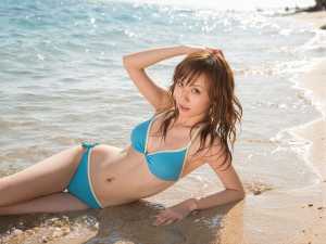 青いビキニを着てビーチでポーズをとり、遊び心のある表情でカメラを直接見つめている若い...。