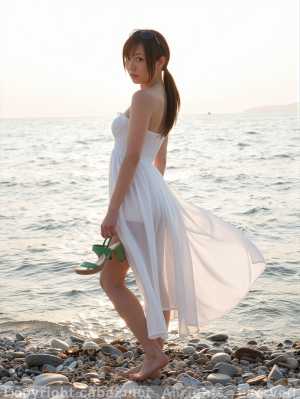 夕暮れ時に小石のビーチに立つ、白いドレスを着た若い女性が緑色のサンダルを履いている様...。