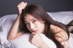 白いシーツが敷かれたベッドに横たわり、思慮深い表情で視聴者をまっすぐに見つめている若...。