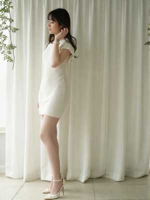 薄手の白いカーテンの前に立つ、白いニットドレスを着た妊婦の横からの写実的な撮影。