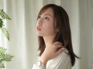 長い茶色の髪を持ち、白いセーターと時計を着て、白いカーテンと緑のシダ植物を背景にした...。