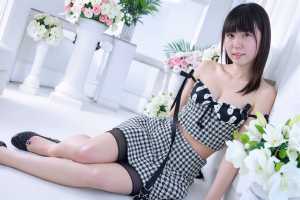 白い部屋で花に囲まれた床に座る若い女性を正面から写実的に撮影した写真。