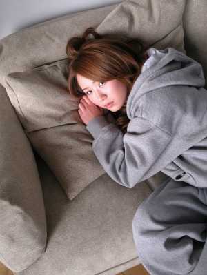 グレーのパーカーを着た若い女性がベージュのソファに座り、思慮深い表情でカメラを直接見...。