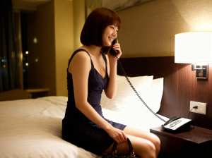 夜のホテルの部屋で、ベッドに座って電話で話している女性を横から写実的に撮影したもので...。