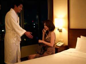 ホテルの一室で、白いバスローブを着た男性が部屋の中央のベッドに座っている女性と握手を...。