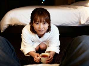 ホテルの部屋のベッドを背景に、白いローブを着た女性がお茶を持って床にしゃがむ様子をロ...。