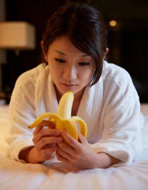 白いバスローブを着た若い女性が、皮をむいたバナナを手に持ってベッドに横たわっている様...。
