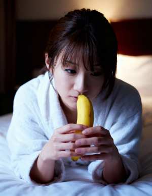 白いバスローブを着てベッドに座り、口の前にバナナを持っている若い女性をクローズアップ...。