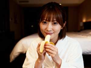ホテルの部屋に座って顔の前にバナナを持っている若い女性を正面カメラのアングルから写実...。