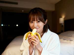 ホテルの部屋のベッドに座って顔の前にバナナを持っている若い女性を、正面カメラのアング...。