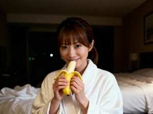 白いローブを着た若い女性がベッドに座り、口の前にバナナを持ってカメラに向かって微笑ん...。