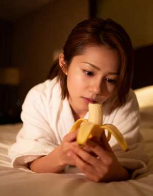 薄暗い寝室の環境でバナナを食べる白いバスローブを着た若い女性を、クローズアップ カメ...。