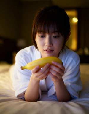 薄暗い寝室のベッドに横たわり、顔の前にバナナを持った若い女性をクローズアップ カメラ...。