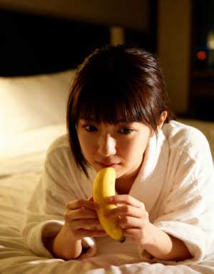 ベッドに座ってバナナを持ち、無表情でカメラを見つめる若い女性を正面から写実的に撮影したもの。