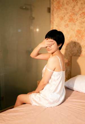 白いシルクのナイトガウンを着て手で目を覆い、ベッドに座っている若い女性を横から撮った...。