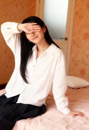 白いボタンダウンシャツと黒いスカートを着て、手で目を覆い、ベッドに座っている若い女性...。