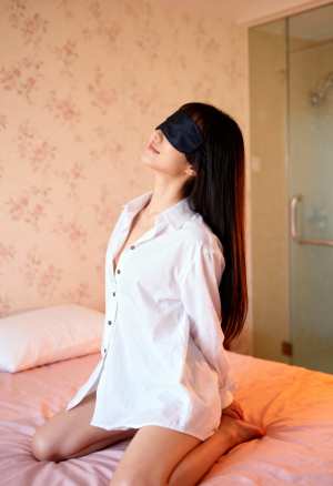 白いボタンダウンのシャツを着て、目隠しをしてベッドに座っている若い女性を横から撮った...。