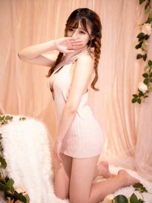 白いバラで飾られた柔らかいピンクの背景の前で、ネックラインが大きく開いたピンクのニッ...。