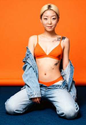 オレンジ色の背景にしゃがみ、オレンジ色のスポーツ ブラとブルー ジーンズを履いた若い...。