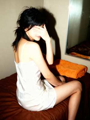 白いスリップドレスを着て手で顔を覆い、ベッドに座っている若い女性を横から撮った写実的な写真。