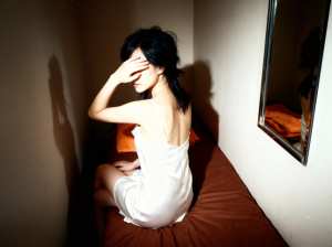 白いスリップドレスを着て、薄暗い部屋のベッドに一人で座っている若い女性を横から撮った...。