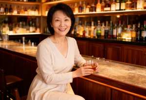 右手にウイスキーのグラスを持ち、バーに座っているアジア系の中年の女性のポートレート。