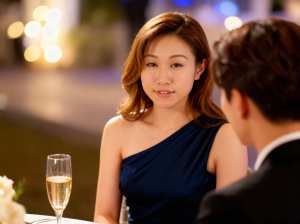 ネイビーブルーのワンショルダードレスを着た若い女性がシャンパングラスを前にテーブルに...。