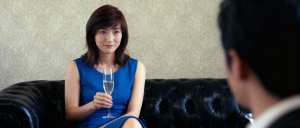 青いドレスを着た女性がシャンパンのグラスを持って黒い革張りのソファに座り、背景の男性...。