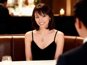 黒いドレスを着て、涙の形のペンダントが付いたネックレスを着て、レストランのテーブルに...。