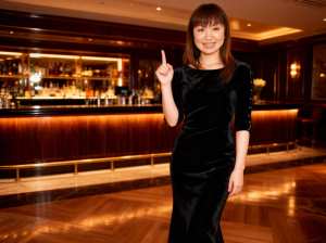 薄暗いバーの真ん中に立ち、自信に満ちた笑顔で親指を立てるジェスチャーをする若い女性。