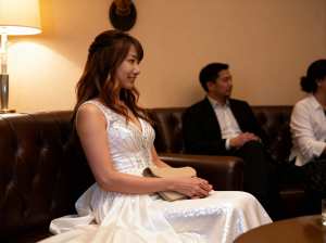 白いウェディングドレスを着た女性が薄暗い部屋で茶色の革張りのソファに座り、笑顔で横を...。