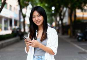 街の通りの真ん中に立ち、スマートフォンを手に持ち、カメラに向かって微笑むアジア系の若い女性。