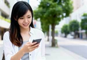 都会のベンチに座り、笑顔で携帯電話を見ているアジア系の若い女性。