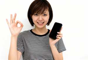 画像の中央に立ち、右手に黒いスマートフォンを持ち、左手でOKサインを出している若い日...。