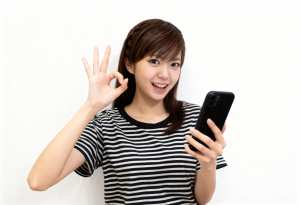 無地の白い背景の前に立ち、右手に黒いスマートフォンを持ち、左手で「OK」のジェスチャ...。