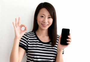 無地の白い背景の前に立ち、右手に黒いスマートフォンを持ち、左手で OK サインを出し...。
