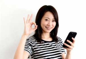 無地の白い背景の前に立ち、右手に黒いスマートフォンを持ち、左手で「OK」のジェスチャ...。