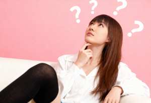 ピンクの背景に白いソファに座り、頭の上に疑問符が浮かび、思慮深い表情で見上げる若い女性。