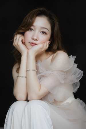 薄手のオフショルダーのトップと繊細なブレスレットを身に着け、あごに手を当てて瞑想的に...。