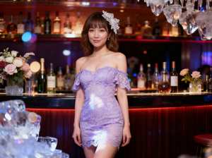 ボトルと花を背景にバーの前に立つ若い女性を正面カメラのアングルから写実的に撮影したもの。