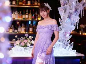 ラベンダーのレースのドレスを着た若い女性がボトルを背景にバーの前に立っている様子を、...。