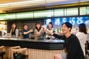 5人の若者がモダンなバーのバーカウンターの周りに集まり、笑顔で飲み物で乾杯した。