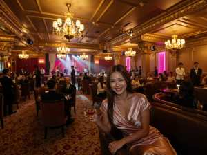 高級レストランに座って赤ワインのグラスを持ち、カメラに向かって微笑む女性を横から写実...。