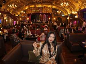 高級レストランで大勢の人々に囲まれ、ビールのグラスを持って座っている女性を正面から写...。