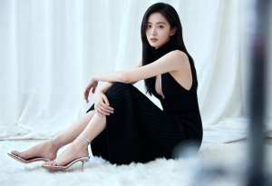 黒いニットのノースリーブドレスと銀のハイヒールを着て、ふわふわの白い敷物の上に座って...。